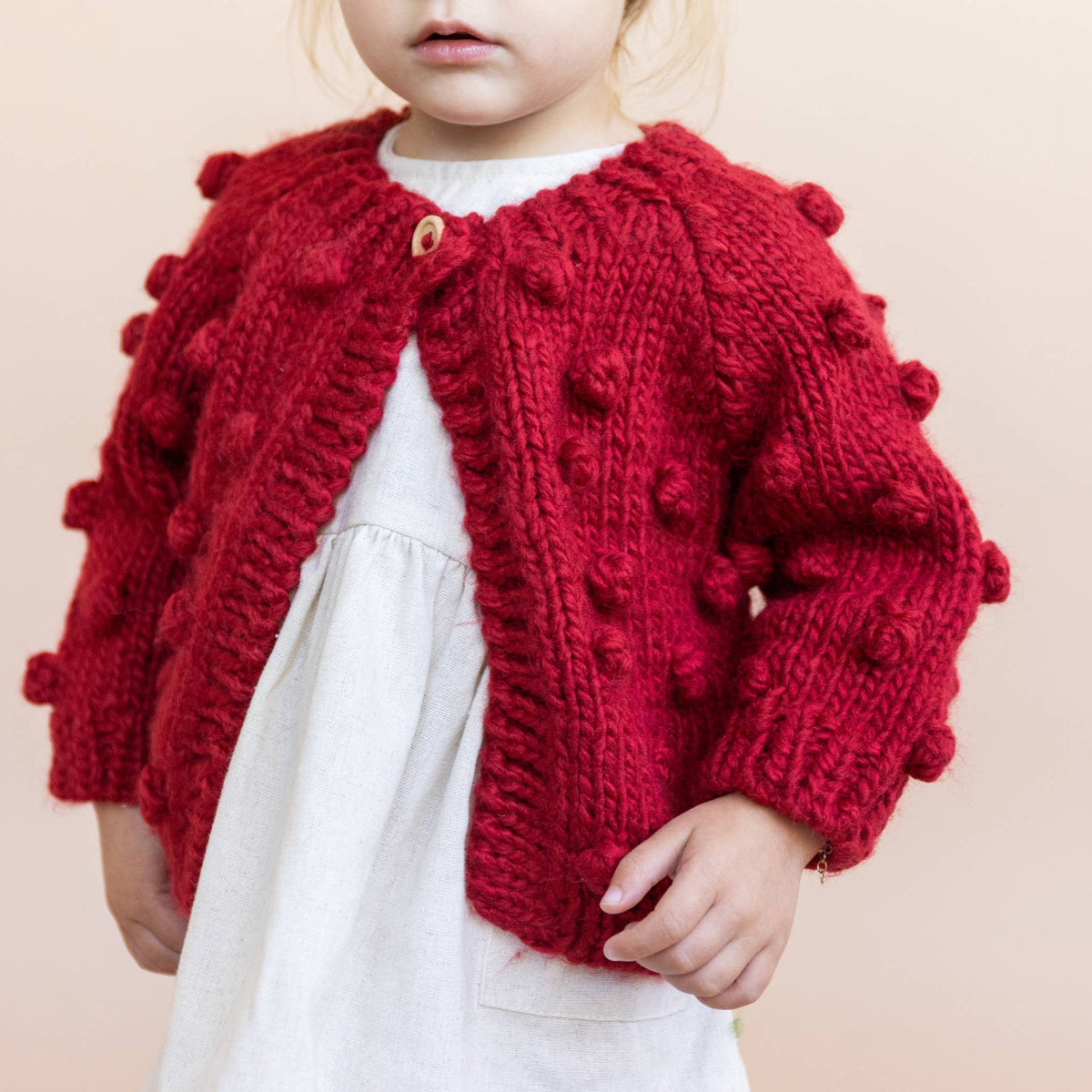 The Blueberry Hill Child wearing the red Popcorn Cardigan over a cream dress